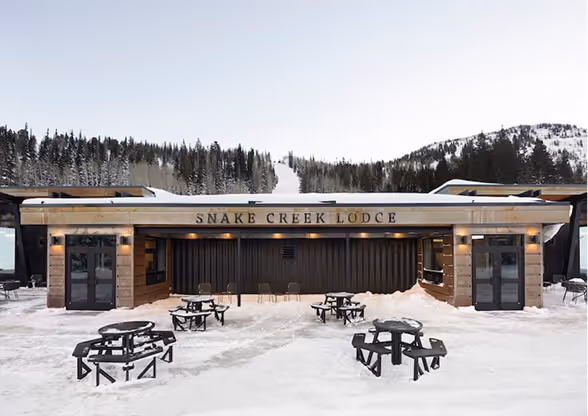 Snow-covered outdoor dining area with round picnic tables in front of Snake Creek Lodge surrounded by forested hills.