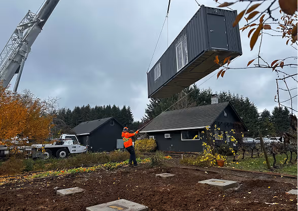 A crane lifts a large gray container above a yard next to a house while a person in an orange jacket guides it with a rope.