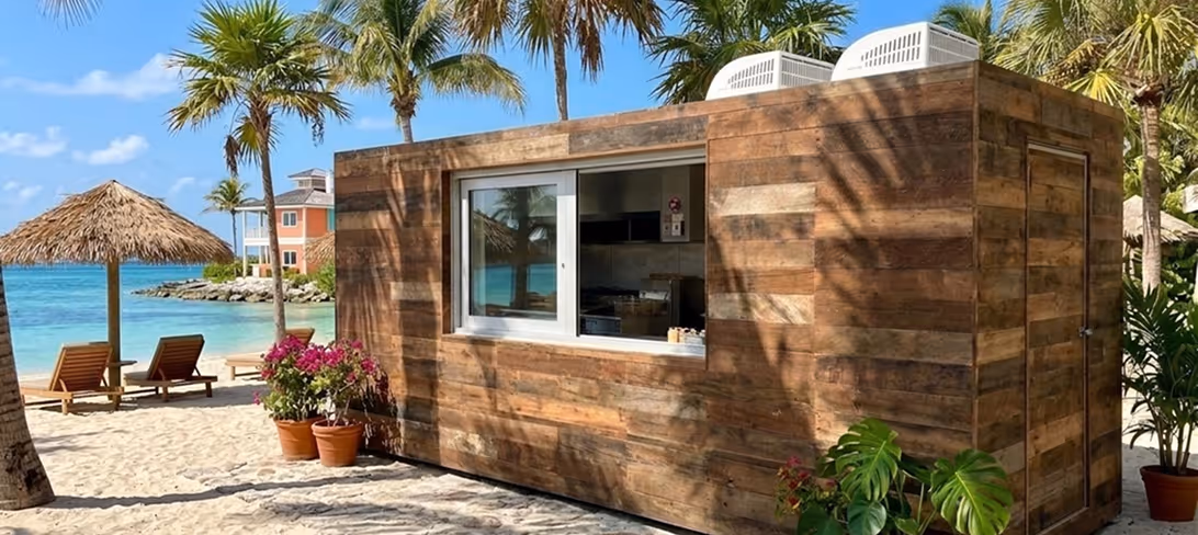 Wooden beach kiosk with a serving window on sandy shore with palm trees and ocean view.