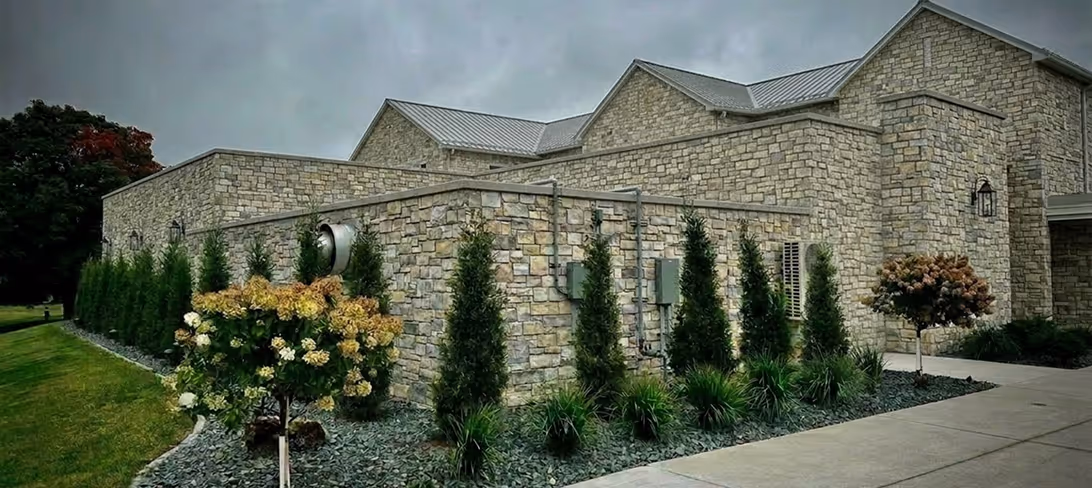 Stone building with multiple roof peaks surrounded by tall shrubs, small trees, and a paved walkway.