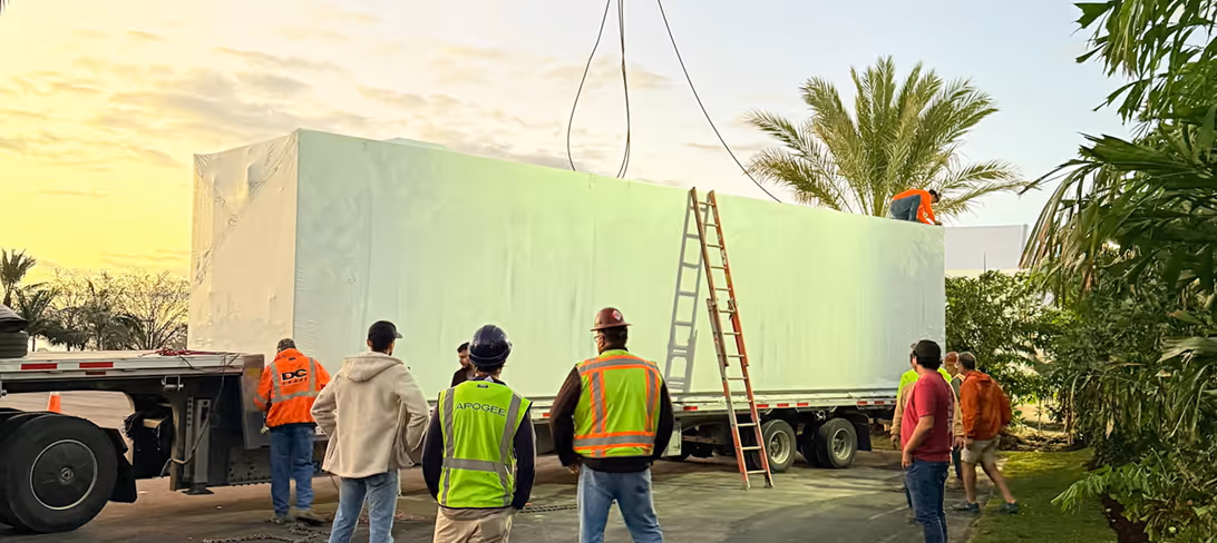 Construction workers guide a large wrapped structure being lifted onto a trailer at sunset.