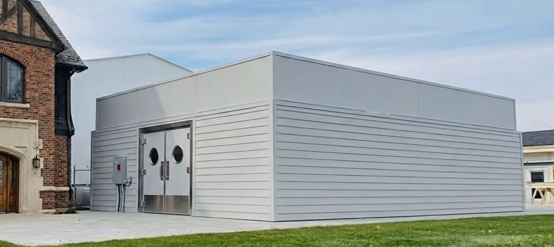 Modern gray utility building with double metal doors and round windows beside a brick building.