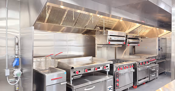 Commercial kitchen with stainless steel appliances including fryers, griddle, ovens, and exhaust hood.