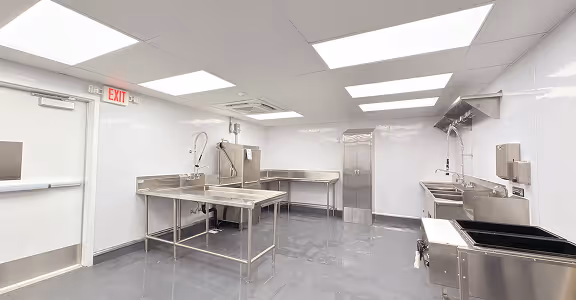 Modern empty commercial kitchen with stainless steel tables, sinks, and bright overhead lighting.