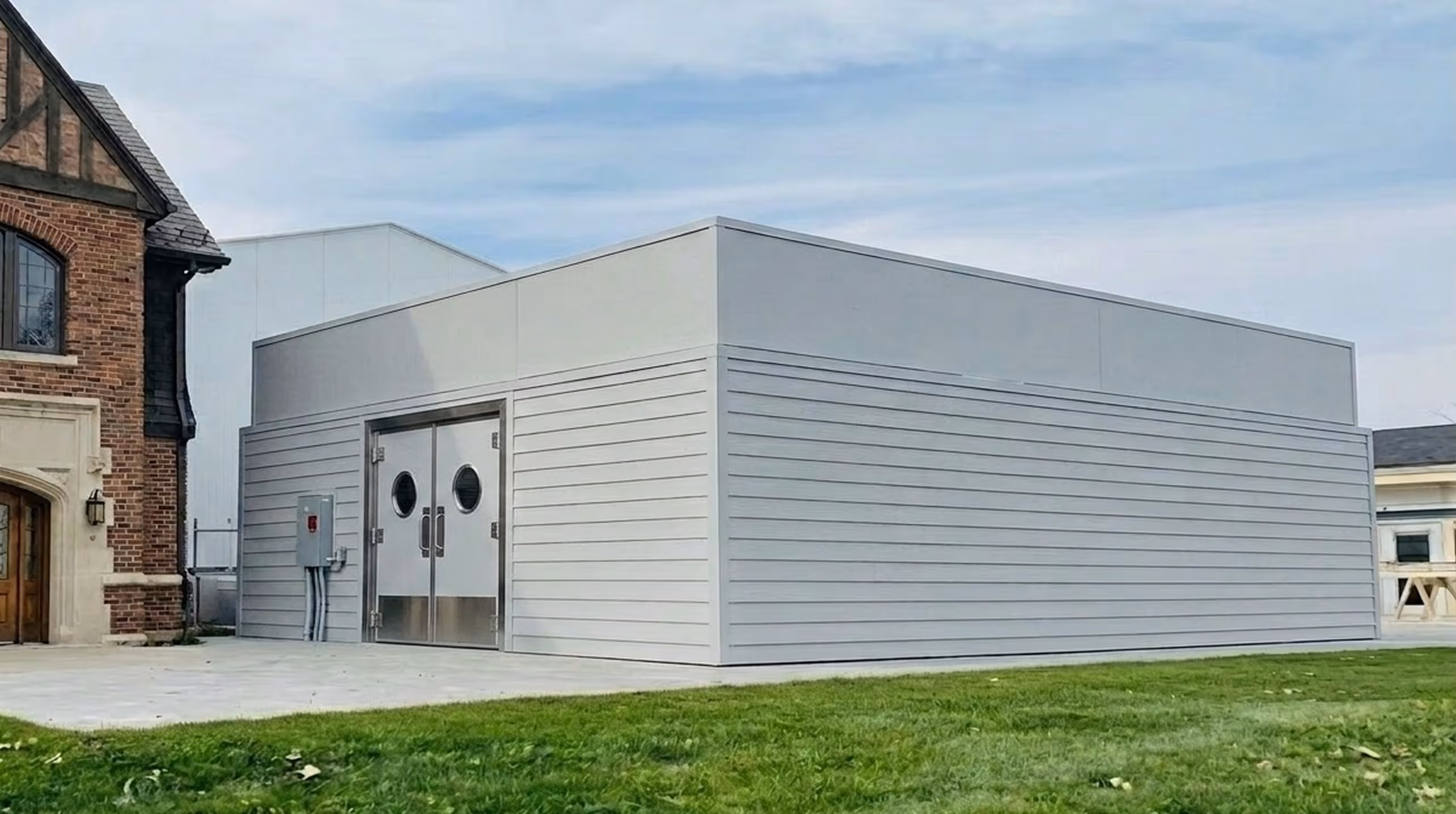 Modern gray modular kitchen building with double metal doors and round windows next to a brick structure on grass.