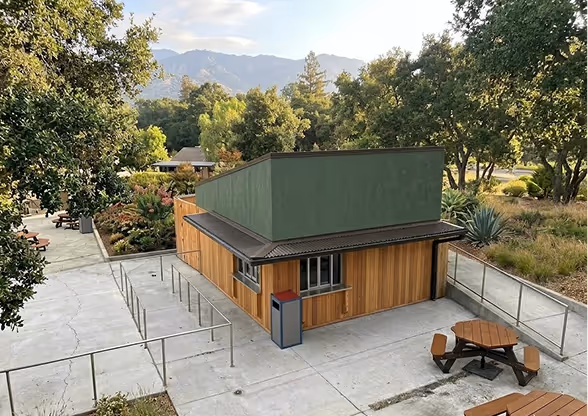 Small wooden modular commercial kitchen with green upper section surrounded by trees, picnic tables, and concrete pathways.