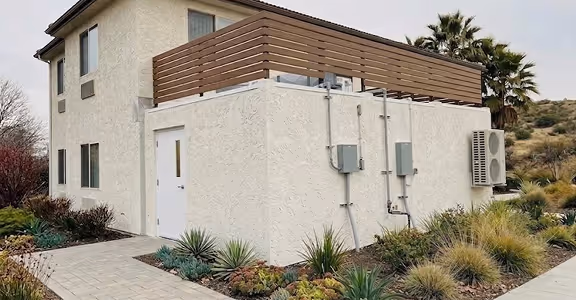 Two-story full service kitchen attached to a house with a wooden balcony railing, air conditioner, and landscaped garden.