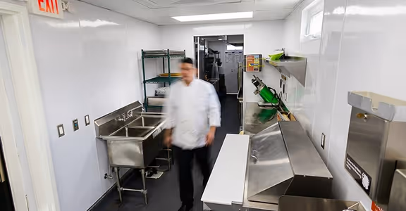 Chef walking in a clean commercial kitchen with stainless steel sinks and counters.