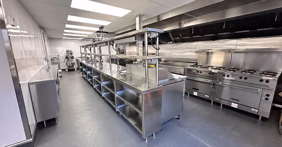 Modern commercial kitchen with stainless steel counters, shelves, and cooking range.