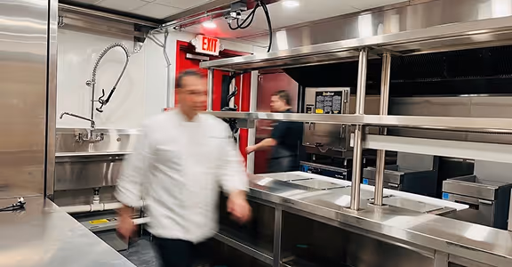 Blurred chef walking in a clean stainless steel quick service commercial kitchen with a red exit door.