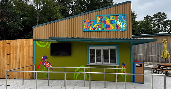 Small green quick service kiosk with colorful bird murals, window, counter, and outdoor seating area.