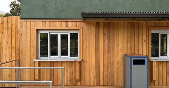 Wood-paneled exterior wall of a modular kitchen with two windows, a metal railing, and a trash bin.
