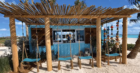 Wooden pergola with blue swings on a sandy beach next to a bar and ocean.