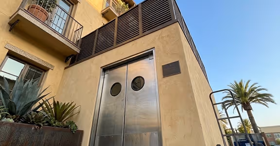 Metal double doors with round windows on a beige commercial kitchen exterior under clear blue sky.