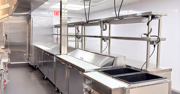Empty modern stainless steel commercial kitchen with prep stations and overhead hanging heat lamps.