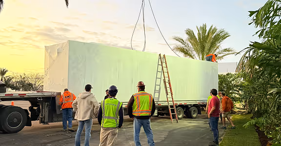 Workers in safety vests guiding a large white module kitchen on a flatbed truck outdoors at sunset.