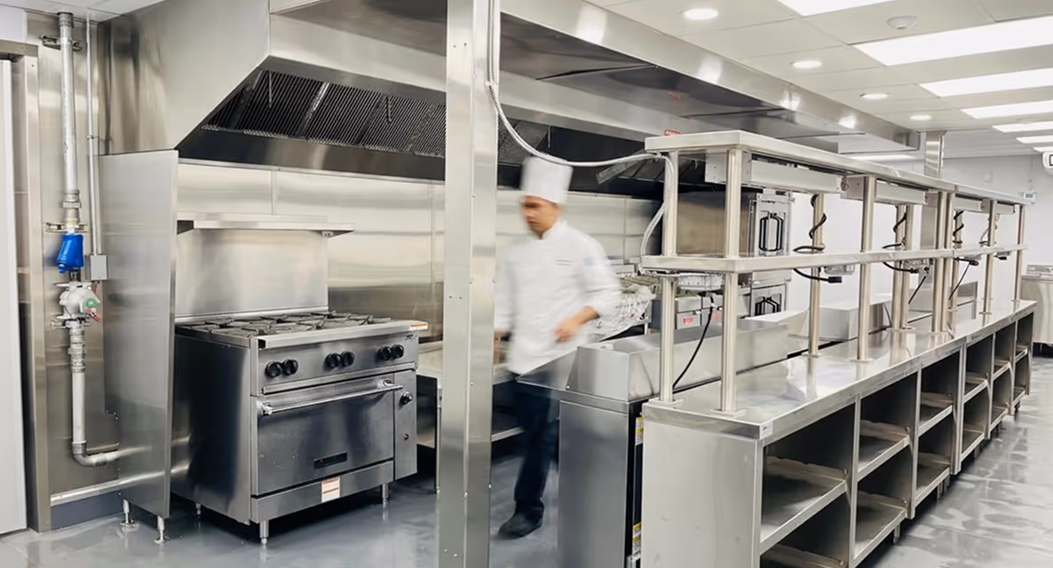 Chef walking in a clean commercial kitchen with stainless steel appliances and counters.