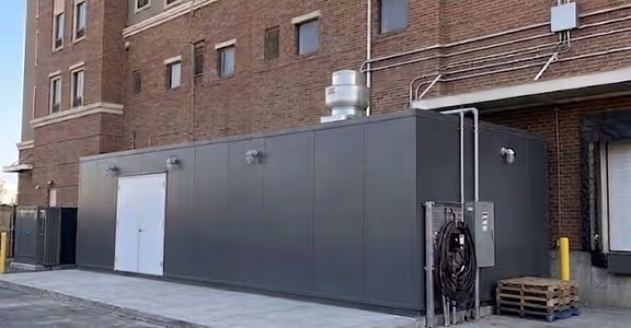 Large gray metal industrial enclosure attached to a brick building with ventilation and cables.
