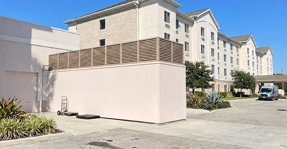 Exterior view of a hotel building with beige walls, plants, a clear sky, and a parked white van.