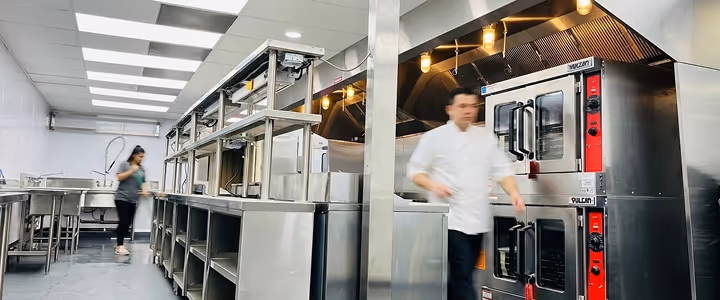 Commercial kitchen with stainless steel appliances and two blurred workers moving inside.