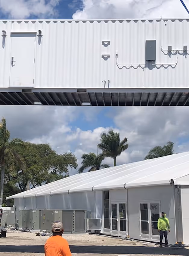 White modular container with door elevated above a large white tent and two people outside.
