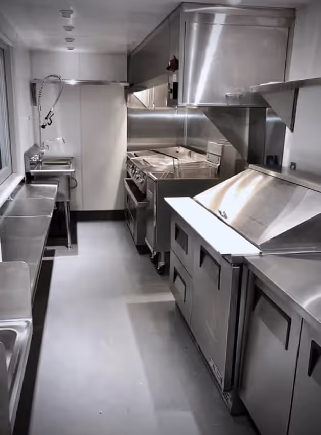 Narrow commercial kitchen with stainless steel appliances, counters, sinks, and stove.