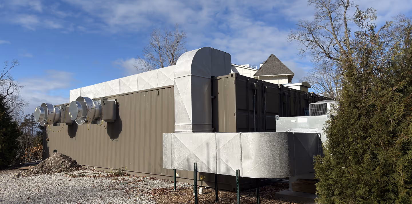 Large brown shipping container with attached metal ventilation ducts outdoors near trees under a partly cloudy sky.