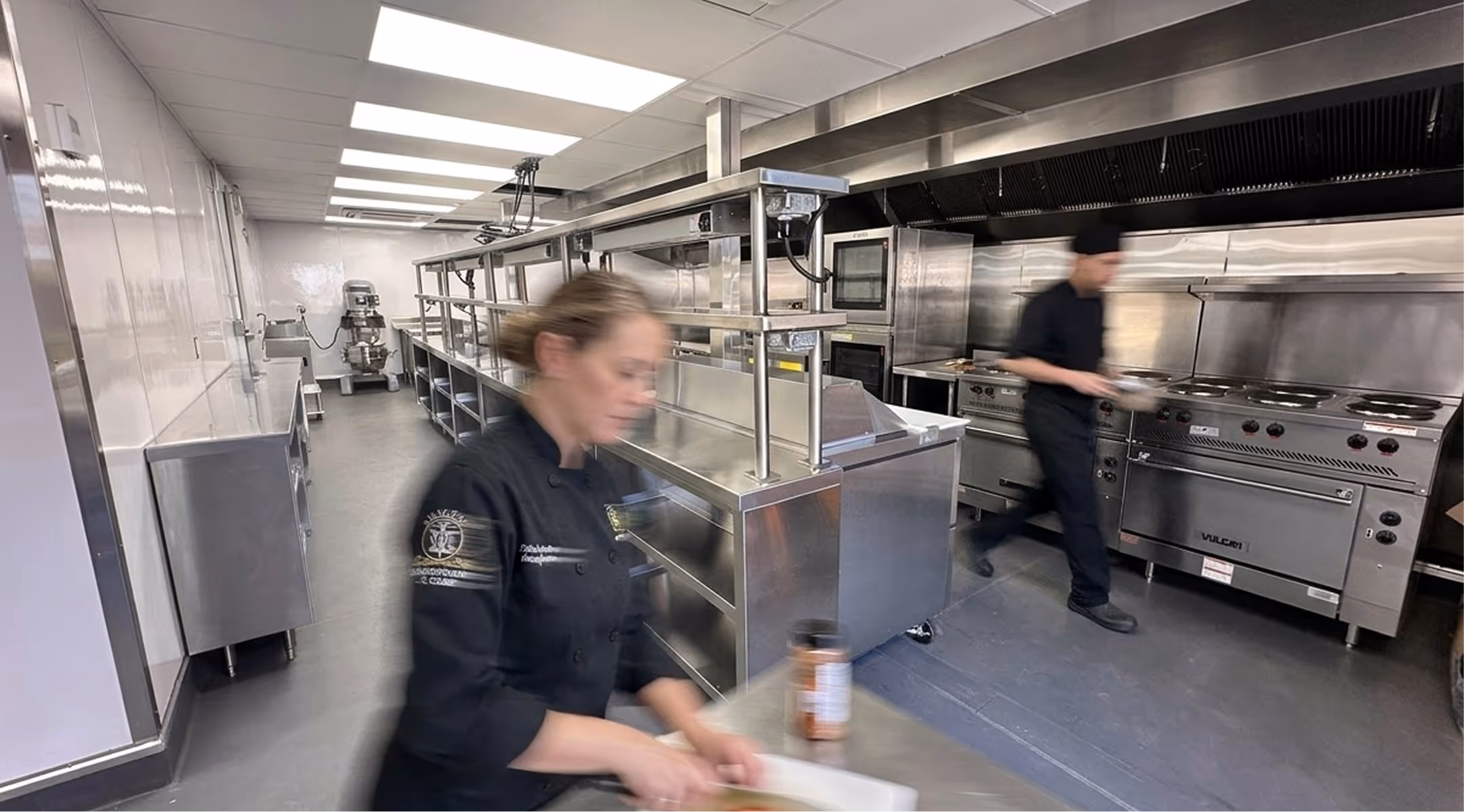 Two blurred chefs working in a clean commercial kitchen with stainless steel appliances and counters.