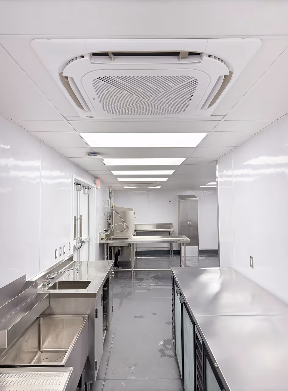 Clean industrial kitchen with stainless steel counters, sinks, and ceiling ventilation unit.