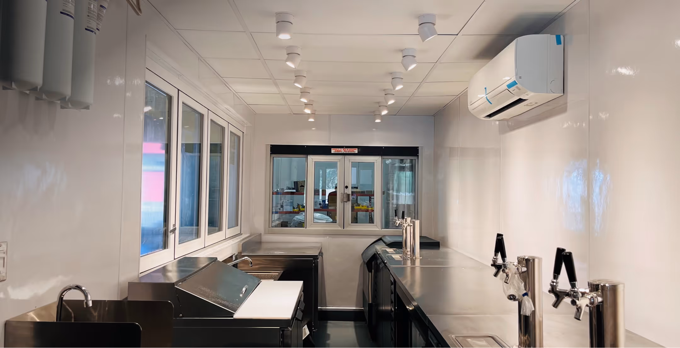 Clean industrial bar kitchen with stainless steel counters, sinks, taps, and overhead lighting.