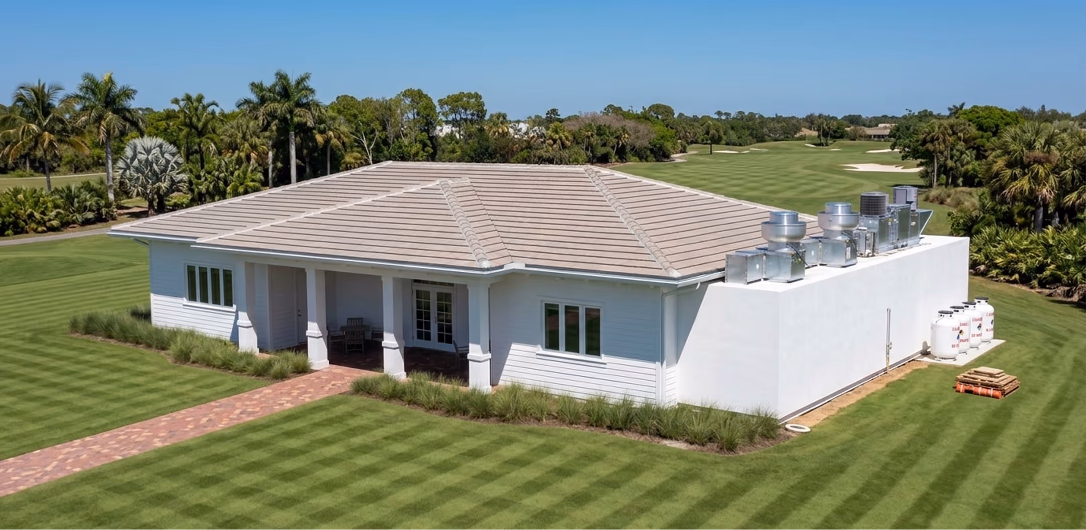 Single-story white building with kitchen and rooftop HVAC units surrounded by manicured grass and palm trees.