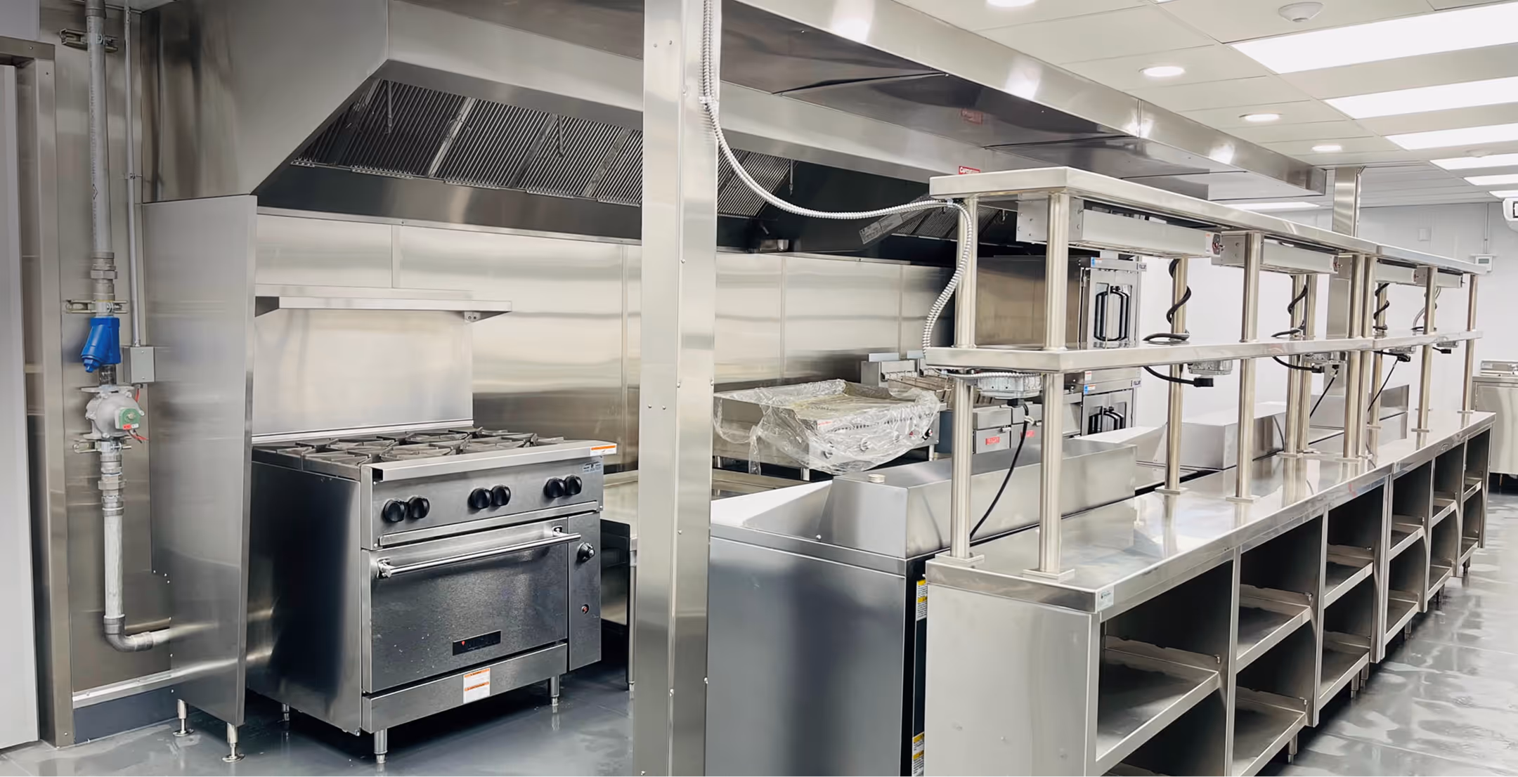 Stainless steel commercial kitchen with oven, stove, prep counters, and a ventilation hood.