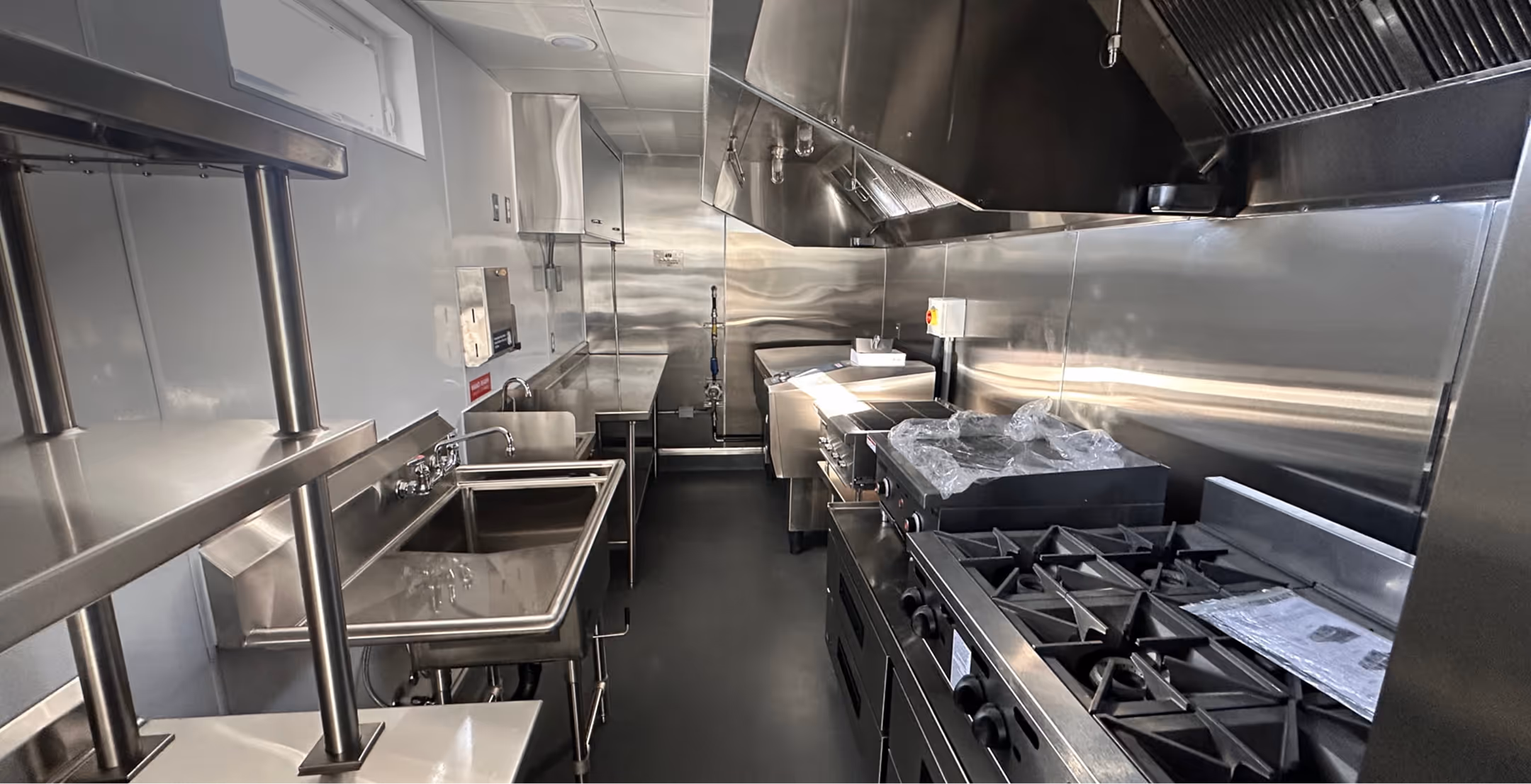 Modern fast food kitchen interior with stainless steel appliances, sinks, stovetop, and ventilation hood.
