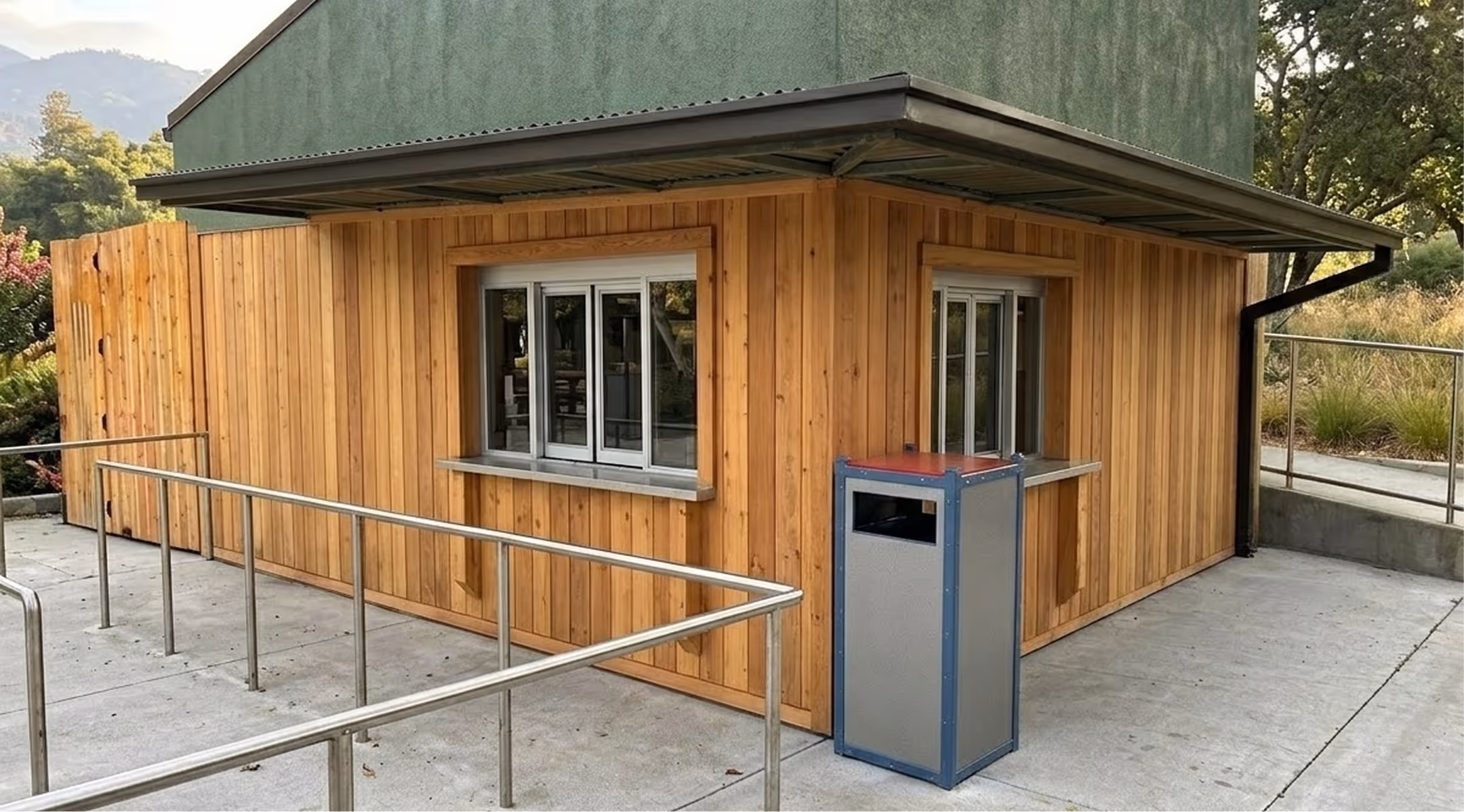 Wood-paneled outdoor kiosk with two service windows, metal railings, and a trash bin on concrete pavement.