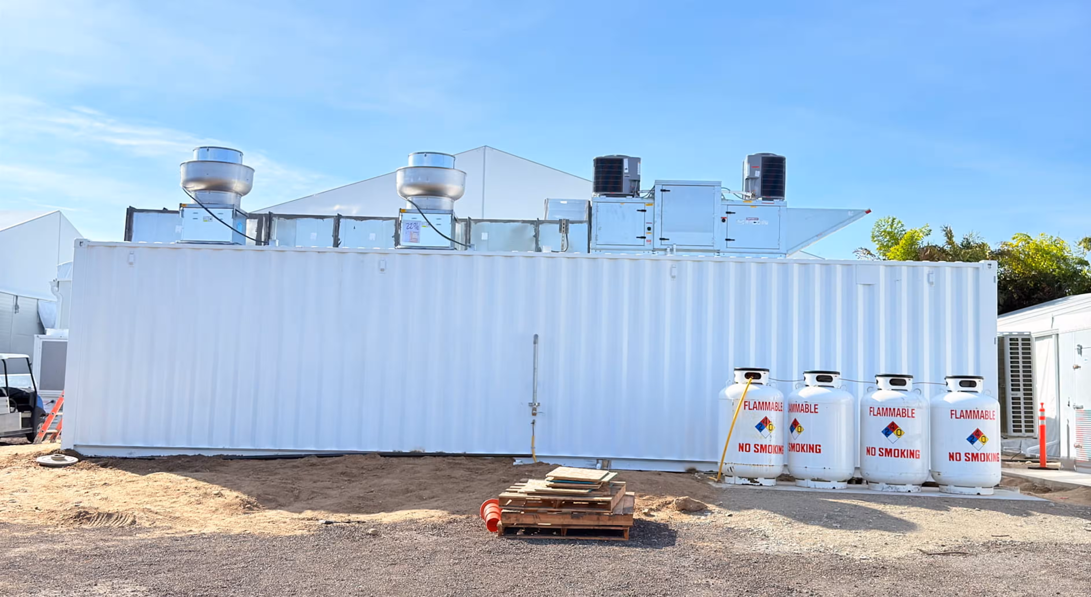 White shipping container kitchen with rooftop ventilation units and four propane tanks labeled flammable no smoking.
