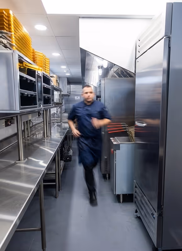 Chef in dark uniform running through a narrow industrial kitchen with stainless steel surfaces.