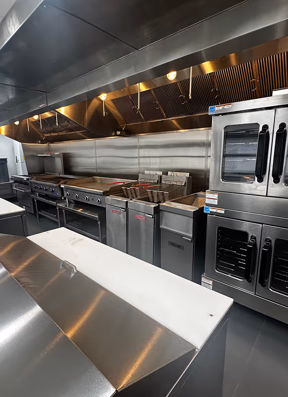 Clean commercial kitchen with stainless steel appliances, fryers, ovens, and white prep counters.