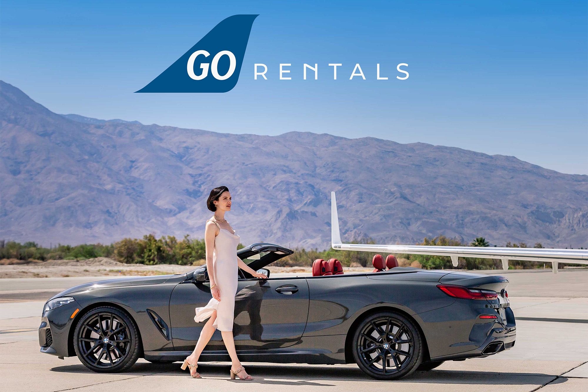 Woman in white dress walking by a black convertible car with mountains in the background. The Go Rentals logo is in the blue sky above the car.