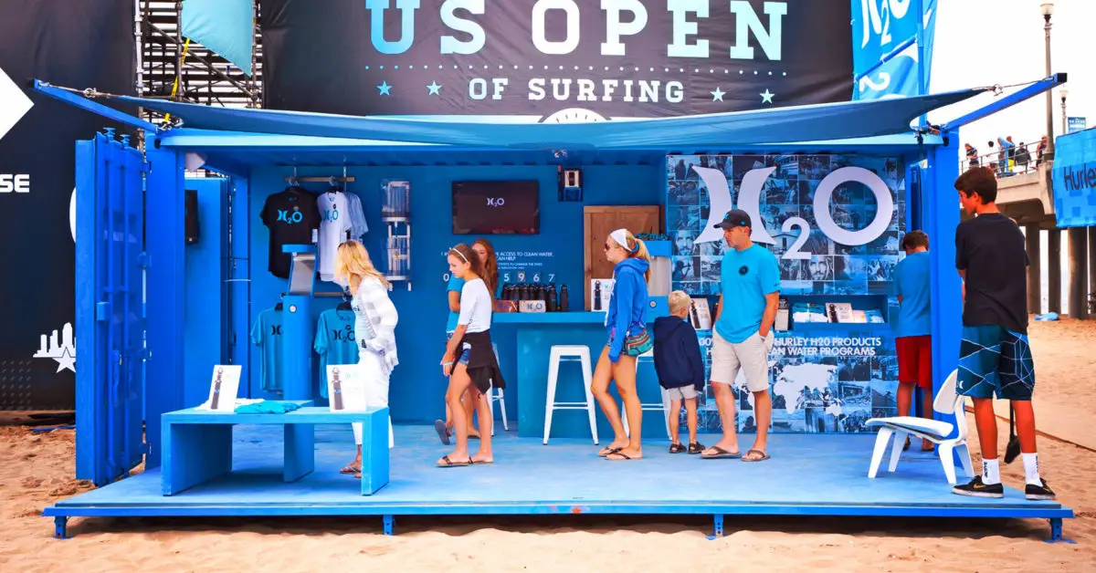 Blue IPME pop-up event container on beach with people browsing H2O water products at US Open of Surfing event