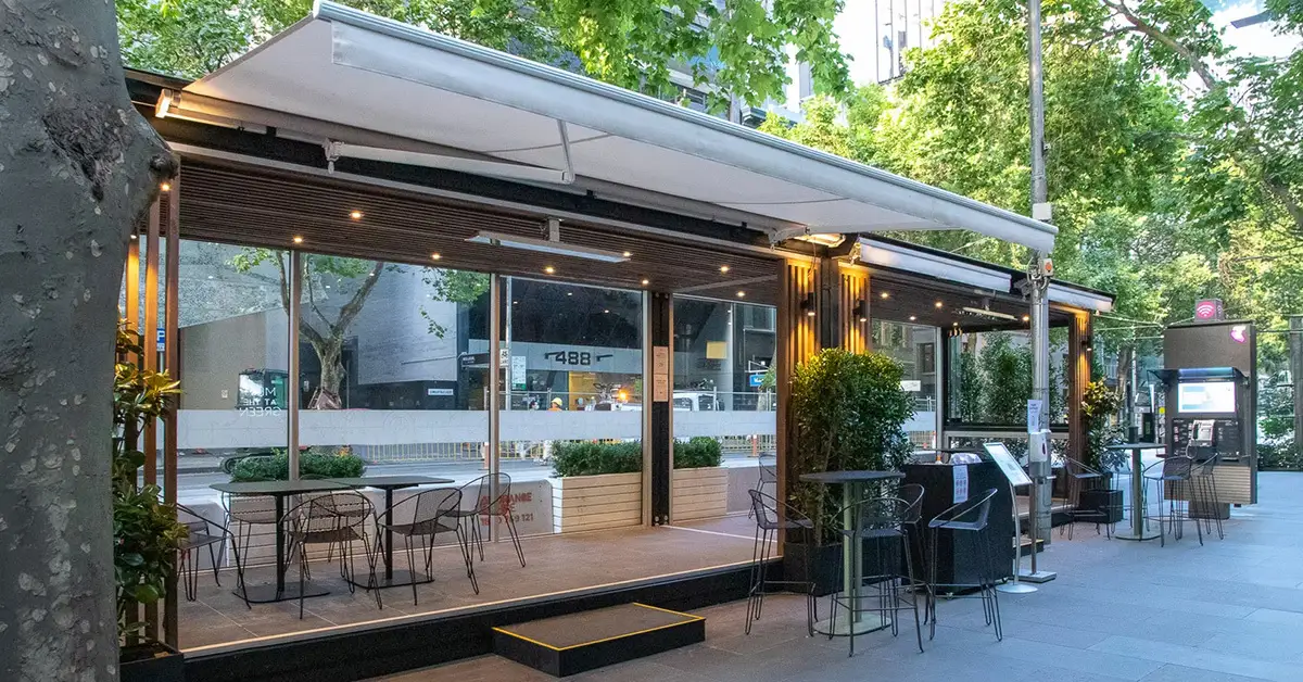 Spacecube expandable contaner set up as a modern outdoor café seating area with metal chairs, tables, plants and overhead lights on city sidewalk.