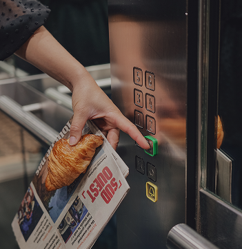 A hand presses an elevator button while holding a croissant and a newspaper.