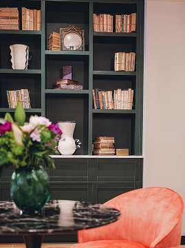 Bibliothèque grise avec livres anciens, objets décoratifs, vase blanc, et bouquet de fleurs sur table ronde noire devant un fauteuil orange.