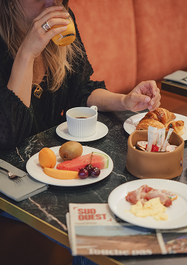 Une personne prenant un verre de jus d’orange avec une table de petit déjeuner comprenant un croissant, une tasse de café, une assiette de fruits et un journal.