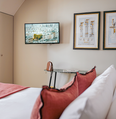 Bed with red cushions, side table with a handbag, wall-mounted television displaying a city view, and two architectural art frames on a beige wall.