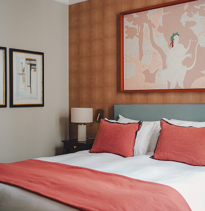 Room with a double bed decorated with cushions and a pink blanket, bedside table with lamp, and framed artwork on the walls.