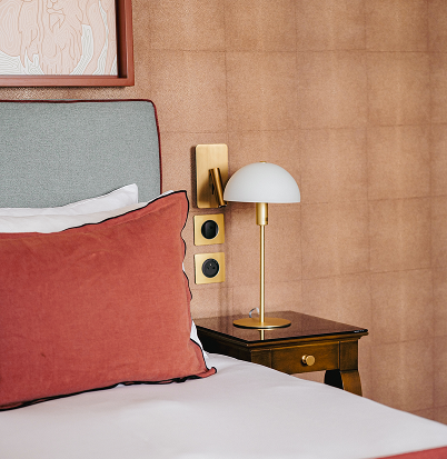 A room with a white bed, a large red cushion, a white and gold lamp on a wooden bedside table, and a beige-colored wall.