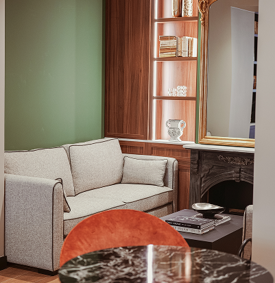 Lounge area with a light grey sofa, a coffee table with books, and a green wall with built-in wooden shelves and a gold-framed mirror.