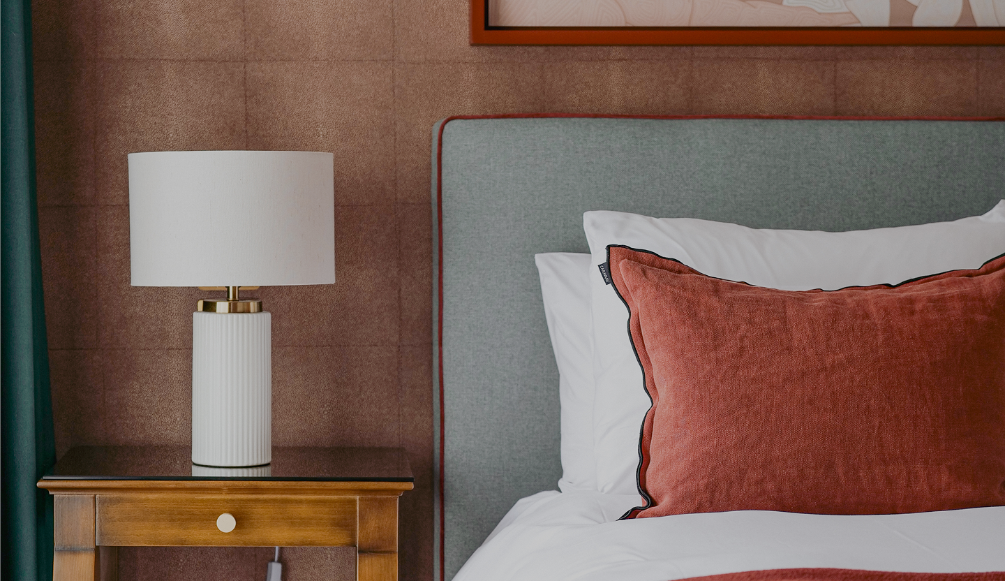 Lampe de table blanche avec base nervurée sur une table de chevet en bois à côté d'un lit avec tête de lit grise et un coussin rouge.
