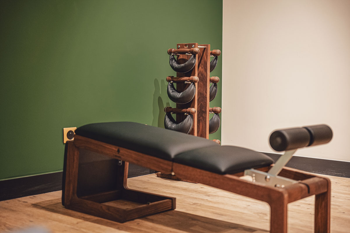 Banc de musculation en bois avec coussins noirs et supports pour haltères contre un mur vert et un mur blanc.