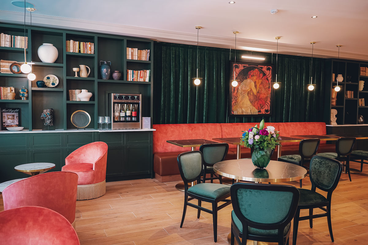 Salle à manger élégante avec fauteuils en velours rose, chaises vert foncé, tables rondes, étagères vert foncé remplies de livres et décorations, et tableau artistique au mur.
